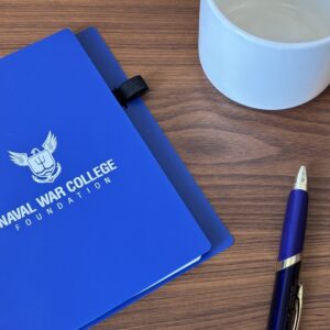Small blue notebook with "Naval War College Foundation" printed on the cover sitting next to a pen and coffee mug on a desk.